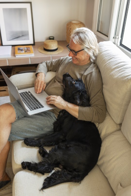 Älterer Mann sitzt mit Hund auf dem Sofa und schaut auf Laptop