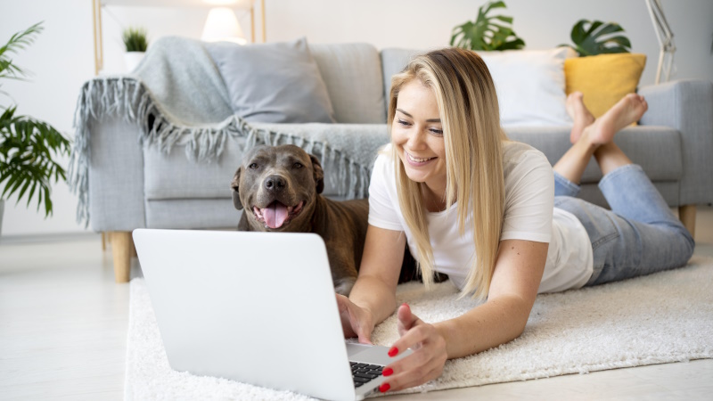 Lächelnde Frau sitzt mit Hund vorm Laptop im Wohnzimmer
